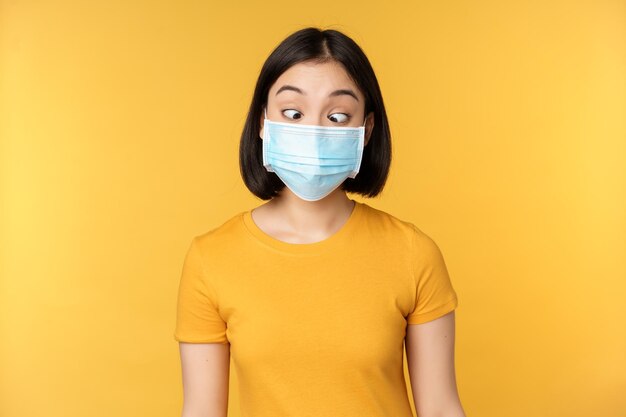 Concetto di salute e persone Divertente ragazza asiatica che strizza gli occhi guardando la sua maschera medica facciale in piedi su sfondo giallo