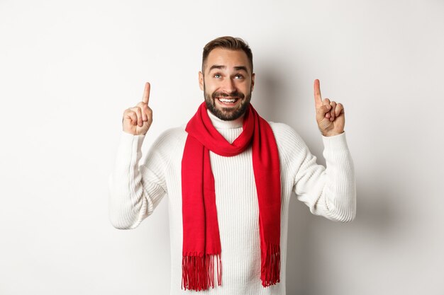 Concetto di shopping natalizio e vacanze invernali. Uomo barbuto felice in sciarpa rossa che punta le dita in alto, guardando eccitato allo spazio della copia, in piedi su sfondo bianco