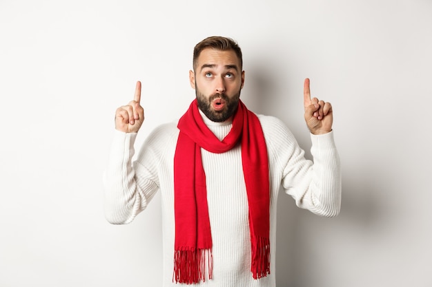 Concetto di shopping natalizio e vacanze invernali. Uomo barbuto stupito che controlla l'offerta promozionale del nuovo anno, indicando e guardando in alto, in piedi su sfondo bianco