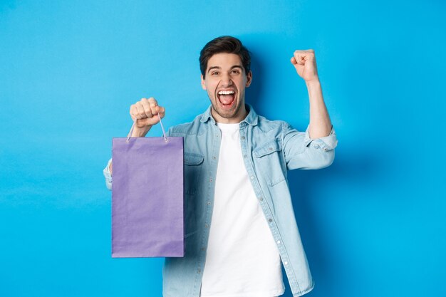 Concetto di shopping, vacanze e stile di vita. Giovane allegro che celebra, tiene in mano un sacchetto di carta e fa una pompa a pugno come un vincitore, in piedi su sfondo blu