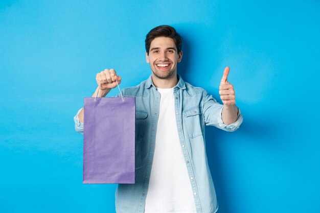 Concetto di shopping, vacanze e stile di vita. Uomo sorridente soddisfatto che tiene in mano un sacchetto di carta, mostra il pollice in su e consiglia il negozio, in piedi su sfondo blu.