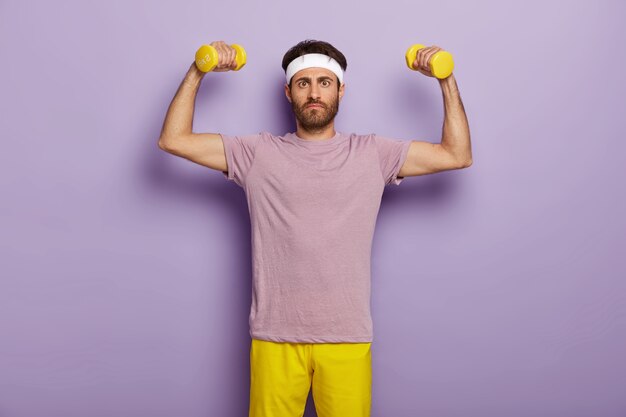 Concetto di sport, esercizio e motivazione. Uomo serio con la barba lunga alza le braccia con manubri, vestito con maglietta viola e pantaloncini gialli, vuole essere sano e forte