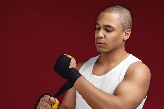 Concetto di sport, forza, potenza e motivazione. Ritratto di bel giovane maschio americano africano kick-boxer preparando i suoi pugni per la lotta, applicando bende di boxe nero, avendo sguardo concentrato