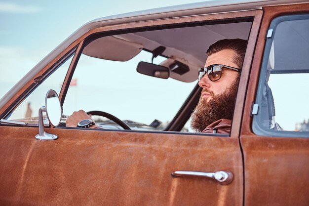 Concetto di stile country. Uomo barbuto in occhiali da sole vestito con giacca di pelle marrone alla guida di un'auto retrò.