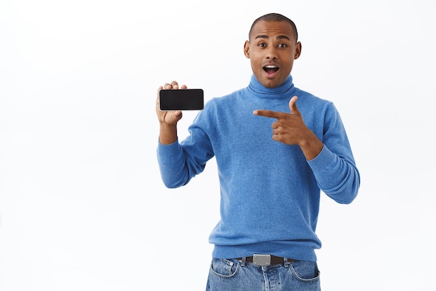 Concetto di stile di vita e persone online su Internet Ritratto di un uomo afroamericano allegro eccitato che parla del nuovo gioco rilasciato per smartphone che punta allo schermo del telefono cellulare sbalordito