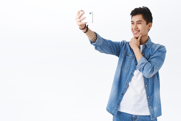 Concetto di stile di vita online di tecnologia Ritratto di uomo asiatico sorridente bello alla moda che prende selfie sullo smartphone tiene il telefono cellulare in mano tesa e cerca di sembrare bello su sfondo bianco della foto