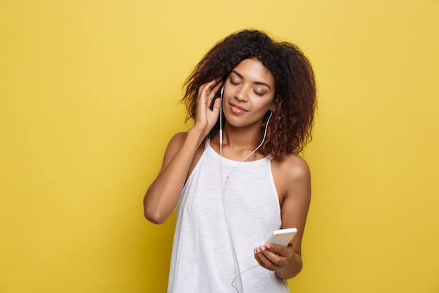 Concetto di stile di vita - Ritratto di bella donna afroamericana allegra ascoltare musica sul cellulare. Sfondo di studio pastello giallo. Copia spazio.