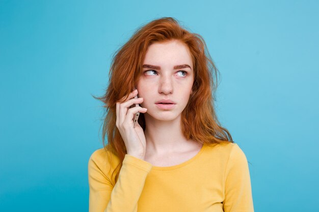 Concetto di stile di vita - Ritratto di ragazza di capelli rossi zenzero con espressione scioccante e stressante mentre parla con l&#39;amico dal telefono cellulare. Isolato su sfondo blu pastello. Copia spazio.