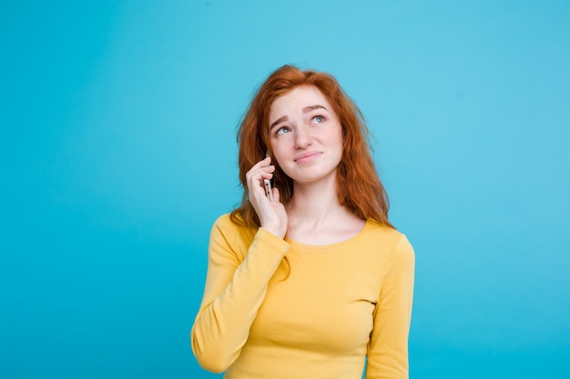 Concetto di stile di vita - Ritratto di ragazza di capelli rossi zenzero con espressione scioccante e stressante mentre parla con l&#39;amico dal telefono cellulare. Isolato su sfondo blu pastello. Copia spazio.