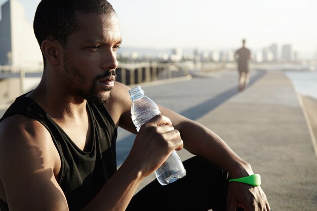 Concetto di stile di vita sano. Ritratto di profilo di sportivo nero con corpo muscoloso seduto sul marciapiede nel sole del mattino dopo esercizi di allenamento, tenendo bottiglia, acqua potabile, guardando in lontananza