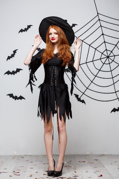 Concetto di strega di Halloween a tutta lunghezza di strega felice di Halloween che tiene in posa su un muro grigio scuro con pipistrello e ragnatela