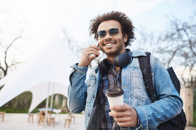 Concetto di tecnologia e persone. Giovane maschio dalla carnagione scura bello con setole e taglio di capelli afro parlando al cellulare mentre beve caffè e cammina per la città, indossando zaino e cappotto di jeans.