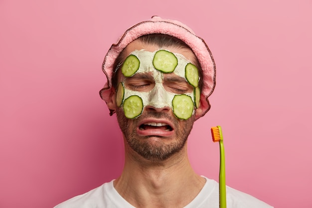 Concetto di trattamento di cura della pelle. L'uomo triste e insoddisfatto guarda tristemente lo spazzolino da denti, ha la barba incolta, applica una maschera cosmetica con i cetrioli