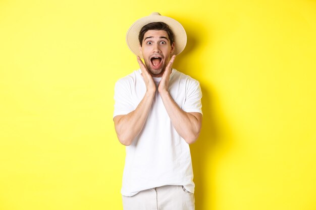 Concetto di turismo ed estate. Sorpreso modello maschile con cappello di paglia, guardando stupito dall'offerta speciale, in piedi su sfondo giallo