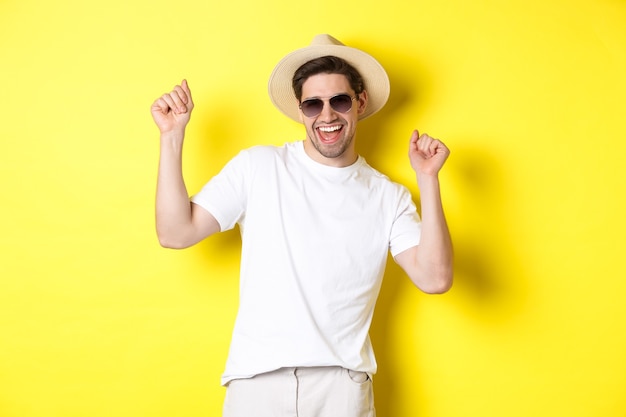 Concetto di turismo, viaggi e vacanze. Felice ragazzo caucasico che balla e si diverte in vacanza, indossando occhiali da sole con cappello di paglia, in piedi su sfondo giallo.