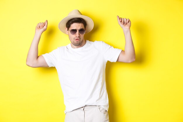 Concetto di turismo, viaggi e vacanze. Turista dell'uomo che si gode le vacanze, balla con cappello di paglia e occhiali da sole, posando su sfondo giallo