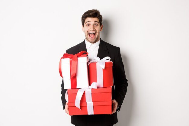 Concetto di vacanze di Natale, celebrazione e stile di vita. Immagine di un bel ragazzo stupito in completo, con in mano regali di capodanno e sorridente, in piedi su sfondo bianco