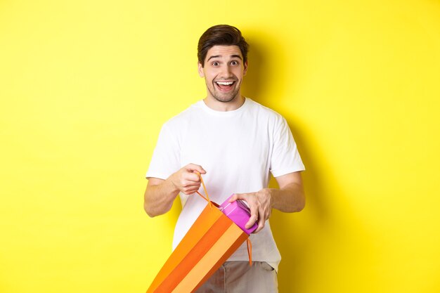 Concetto di vacanze e celebrazione. Uomo felice e sorpreso togliere il regalo e guardando la telecamera, in piedi su sfondo giallo.