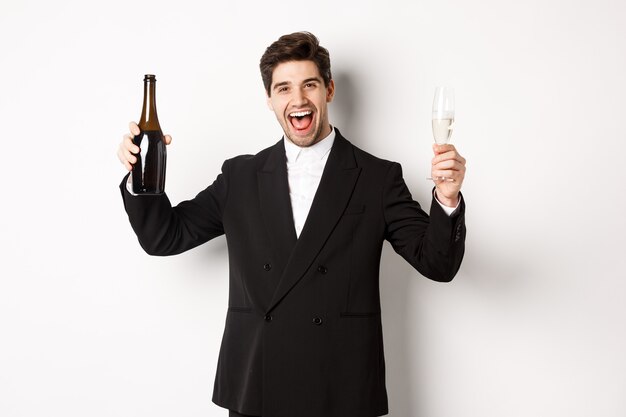Concetto di vacanze, festa e celebrazione. Bell'uomo in abito alla moda divertendosi, tenendo in mano una bottiglia e un bicchiere di champagne, in piedi su sfondo bianco.