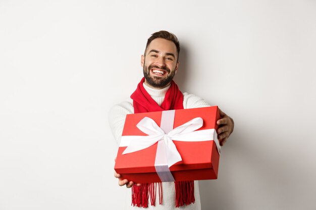 Concetto di vacanze invernali. Bel ragazzo che fa un regalo, augurando buon natale e felice anno nuovo, in piedi su sfondo bianco