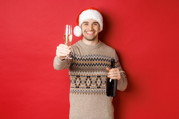 Concetto di vacanze invernali, capodanno e celebrazioni. Ritratto di un bell'uomo con cappello e maglione da babbo natale, con in mano champagne, alzando il bicchiere e salutando la festa di natale