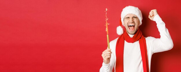 Concetto di vacanze invernali di Natale e celebrazione Uomo bello che festeggia il nuovo anno e si diverte a tenere lo sparkler e sorridente indossando il cappello di Babbo Natale in piedi su sfondo rosso