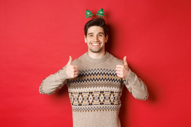 Concetto di vacanze invernali, natale e celebrazione. Ragazzo barbuto allegro in maglione, che mostra il pollice in su in segno di approvazione e sorridente, godendosi la festa di capodanno, sfondo rosso