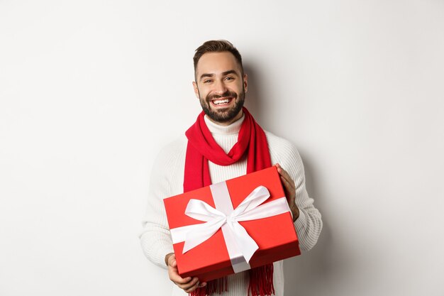 Concetto di vacanze invernali. Uomo sorridente che ti fa un regalo, tiene un regalo e augura buon natale, in piedi su sfondo bianco