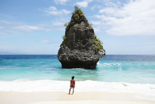 Concetto di viaggio, avventura, hobby e vacanza. Giovane uomo vestito casualmente con cappello nero che cammina lungo la spiaggia sabbiosa deserta, di fronte all'oceano turchese con un'isola di pietra con alte rocce nel mezzo