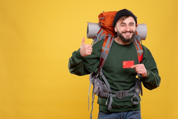 Concetto di viaggio con il giovane ragazzo sorridente con packpack e mostrando la carta di credito che fa il gesto giusto sul giallo