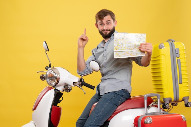 Concetto di viaggio con il giovane uomo barbuto emotivo seduto sulla moto e rivolto in avanti tenendo la mappa rivolta verso l'alto su di esso su giallo