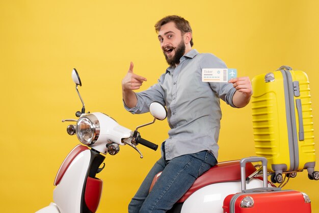 Concetto di viaggio con il giovane uomo barbuto sorridente che si siede sulla motocicletta e che indica il biglietto su di esso su giallo