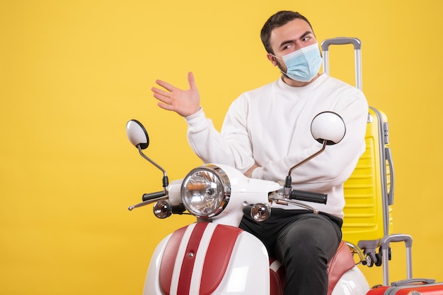 Concetto di viaggio con ragazzo confuso in maschera medica seduto su moto con valigia gialla su di esso su giallo