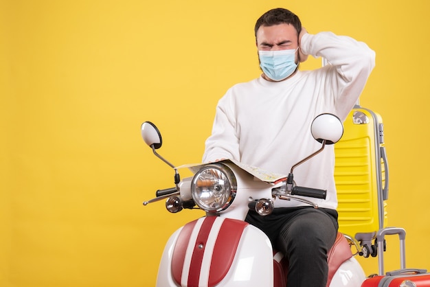 Concetto di viaggio con ragazzo nervoso in maschera medica seduto su moto con valigia gialla su di esso e tenendo la mappa che soffre di mal di testa