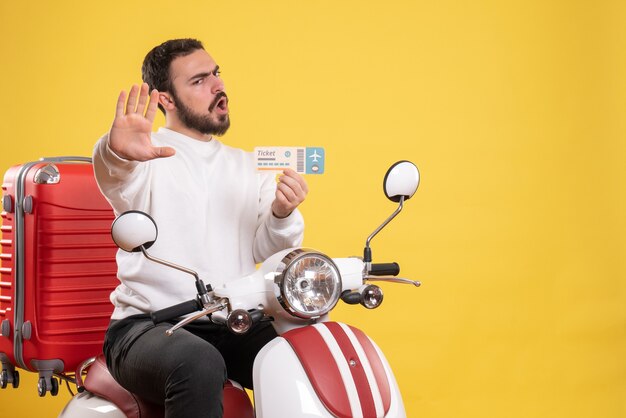 Concetto di viaggio con un giovane uomo emotivo seduto su una moto con la valigia sopra che mostra il biglietto su giallo
