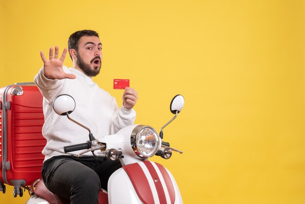 Concetto di viaggio con un giovane uomo in viaggio spaventato seduto su una moto con la valigia sopra che tiene una carta bancaria su giallo