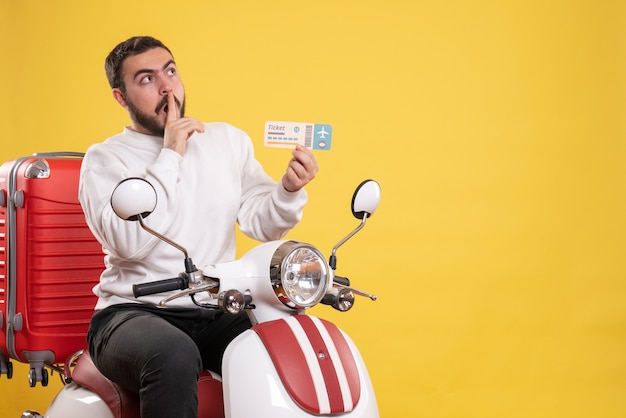 Concetto di viaggio con un uomo in viaggio seduto su una motocicletta con la valigia sopra che mostra il biglietto e alza lo sguardo facendo gesto di silenzio su giallo