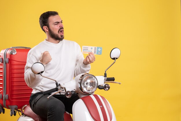 Concetto di viaggio con un uomo sorpreso seduto su una moto con la valigia sopra che mostra il biglietto su giallo