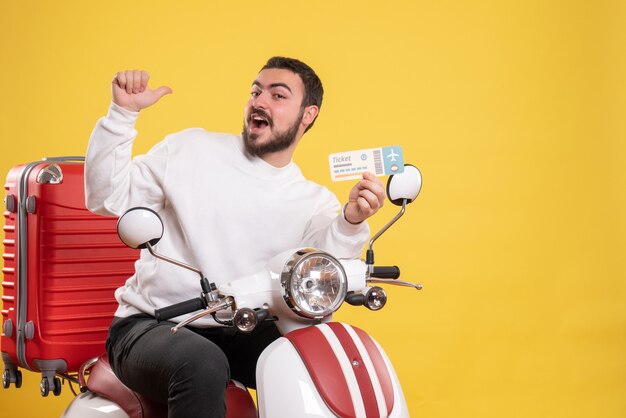 Concetto di viaggio con un uomo sorridente seduto su una moto con la valigia sopra che mostra il biglietto su giallo