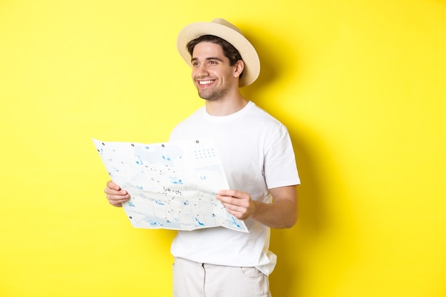 Concetto di viaggio, vacanza e turismo. Bel ragazzo turistico andando in giro per la città, tenendo la mappa e sorridente, in piedi su sfondo giallo.