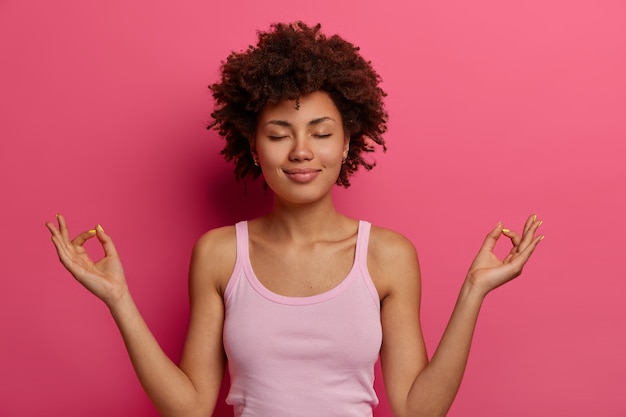 Concetto di yoga e meditazione. La donna dalla pelle scura soddisfatta e rilassata tiene le mani nel gesto del mudra, si sente in pace dopo una dura giornata, tiene gli occhi chiusi, controlla i suoi sentimenti, si trova nella posa del loto.