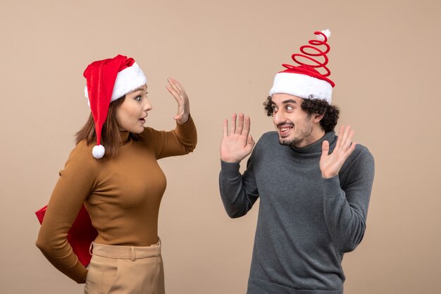 Concetto festivo di umore del nuovo anno con coppia adorabile divertente che porta il regalo nascosto della ragazza dei cappelli di Babbo Natale rosso dietro su gray