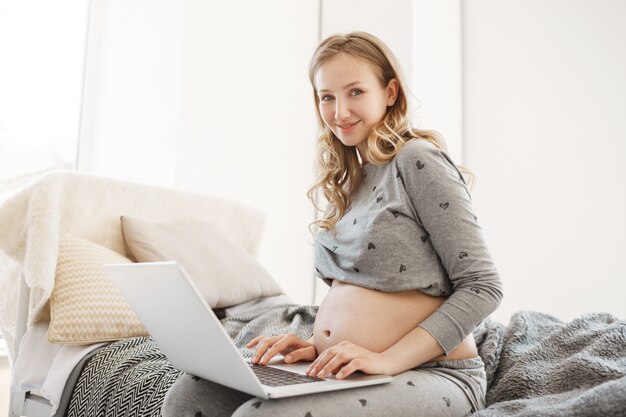 Concetto freelance. Ritratto di allegra giovane donna incinta con i capelli biondi in comodo abbigliamento da casa seduto sul letto lavorando sul computer portatile, creando un nuovo progetto anche durante il periodo di gravidanza