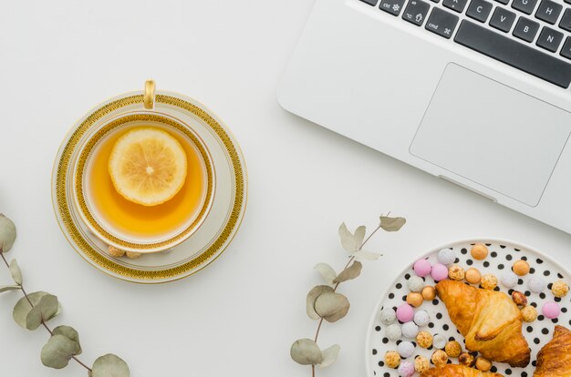 Confetteria e croissant sul piatto con tè al limone vicino al portatile aperto su sfondo bianco