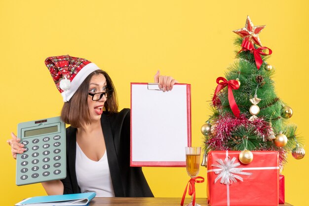 Confusa affascinante signora in vestito con il cappello di Babbo Natale che mostra il documento e tenendo la calcolatrice in ufficio su giallo isolato