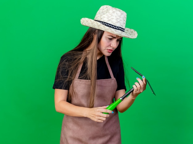 Confusa bella ragazza giardiniere in uniforme che indossa cappello da giardinaggio tenendo e guardando zappa rastrello isolato su sfondo verde