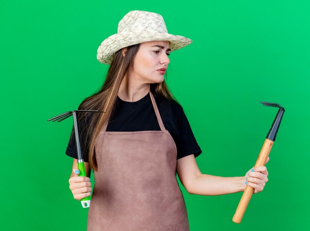 Confusa bella ragazza giardiniere in uniforme che indossa cappello da giardinaggio tenendo la zappa rastrello e guardando il rastrello in mano isolato su sfondo verde