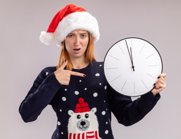 Confusa giovane bella ragazza che indossa il cappello di Natale che tiene e punti all'orologio di parete isolato su priorità bassa bianca