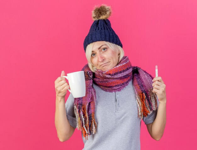 Confusa giovane bionda malata donna slava che indossa il cappello invernale e la sciarpa tiene la tazza