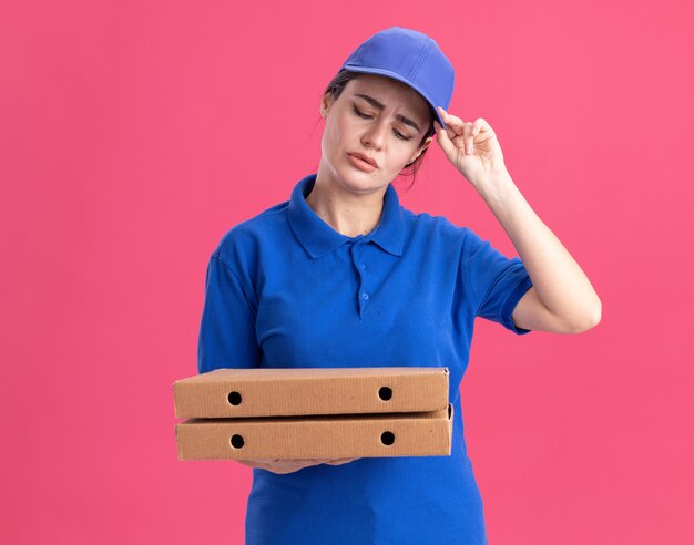 Confusa giovane donna delle consegne in uniforme e berretto che tiene e guarda i pacchetti di pizza che afferrano il berretto